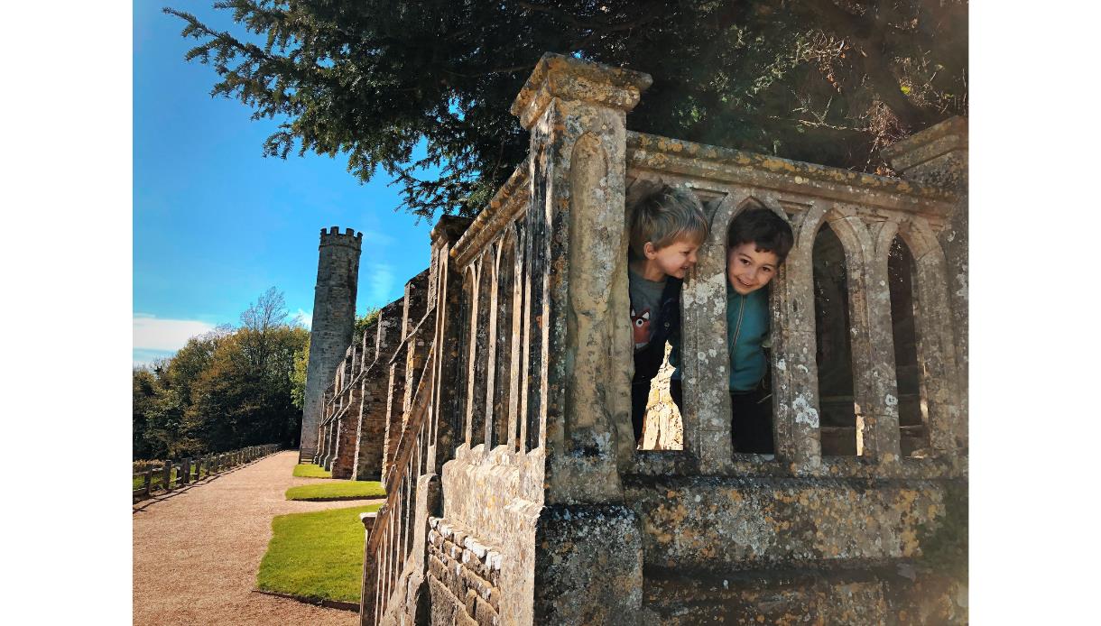 Children peeking through the stone balustrade at Battle Abbey, with historic walls and a tower along a sunny pathway.