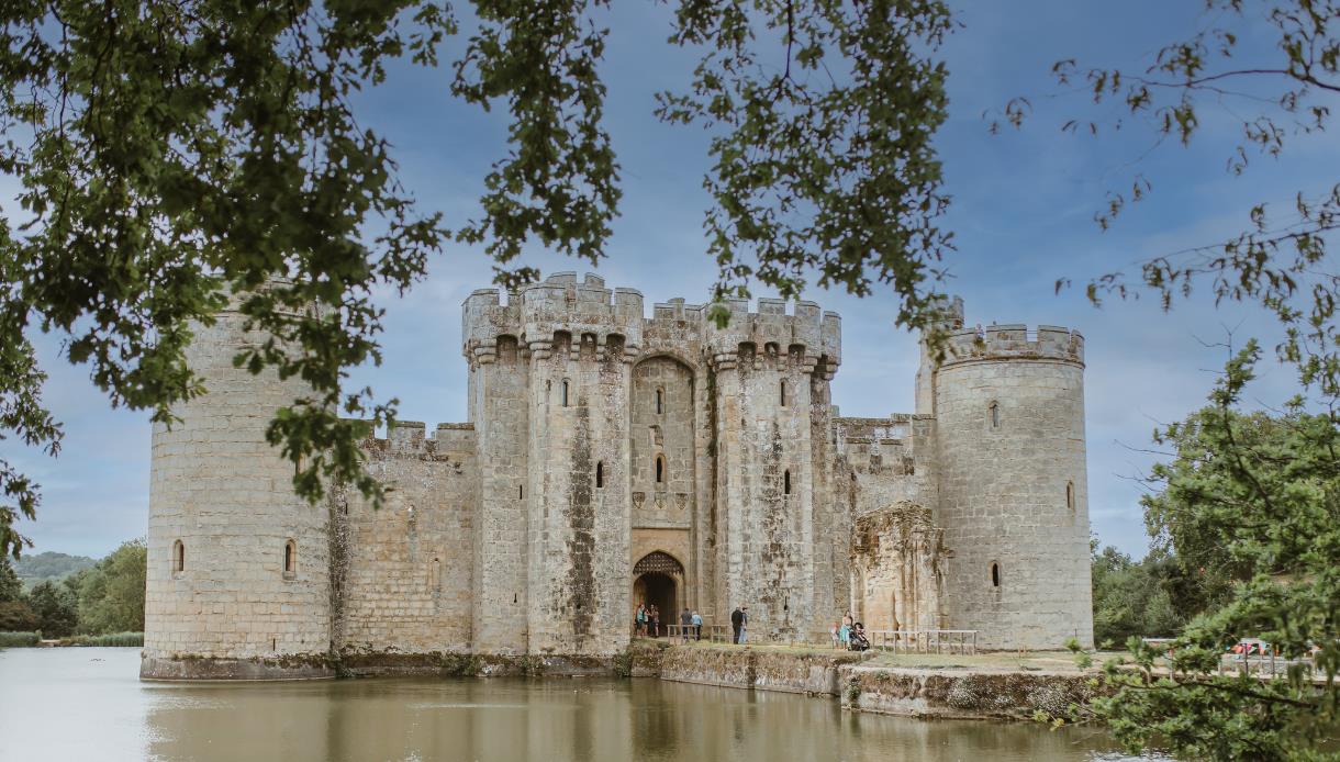 Bodiam Castle reflected in the surrounding moat, framed by tree branches under a bright sky.
