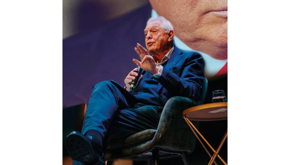 Speaker (John Simpson) on stage in armchair holding microphone , with large screen backdrop and table beside them.
