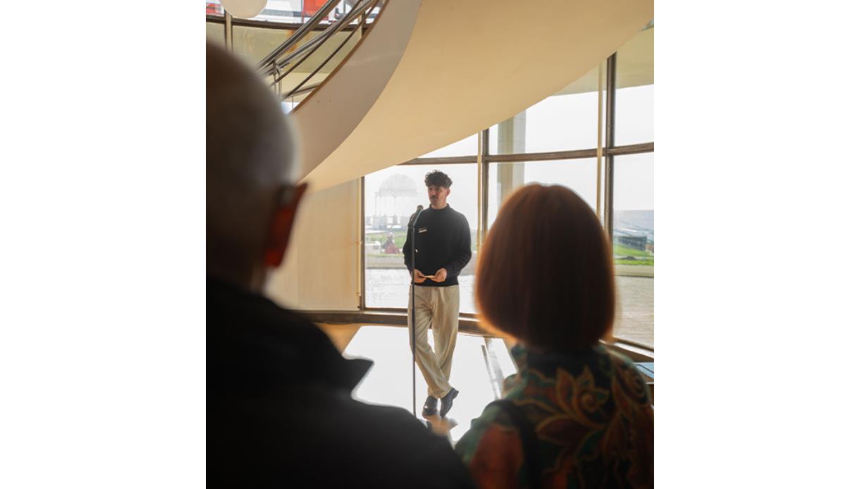 Exhibition tour at De La Warr Pavilion, with a speaker addressing visitors beside the pavilion’s curved window wall.