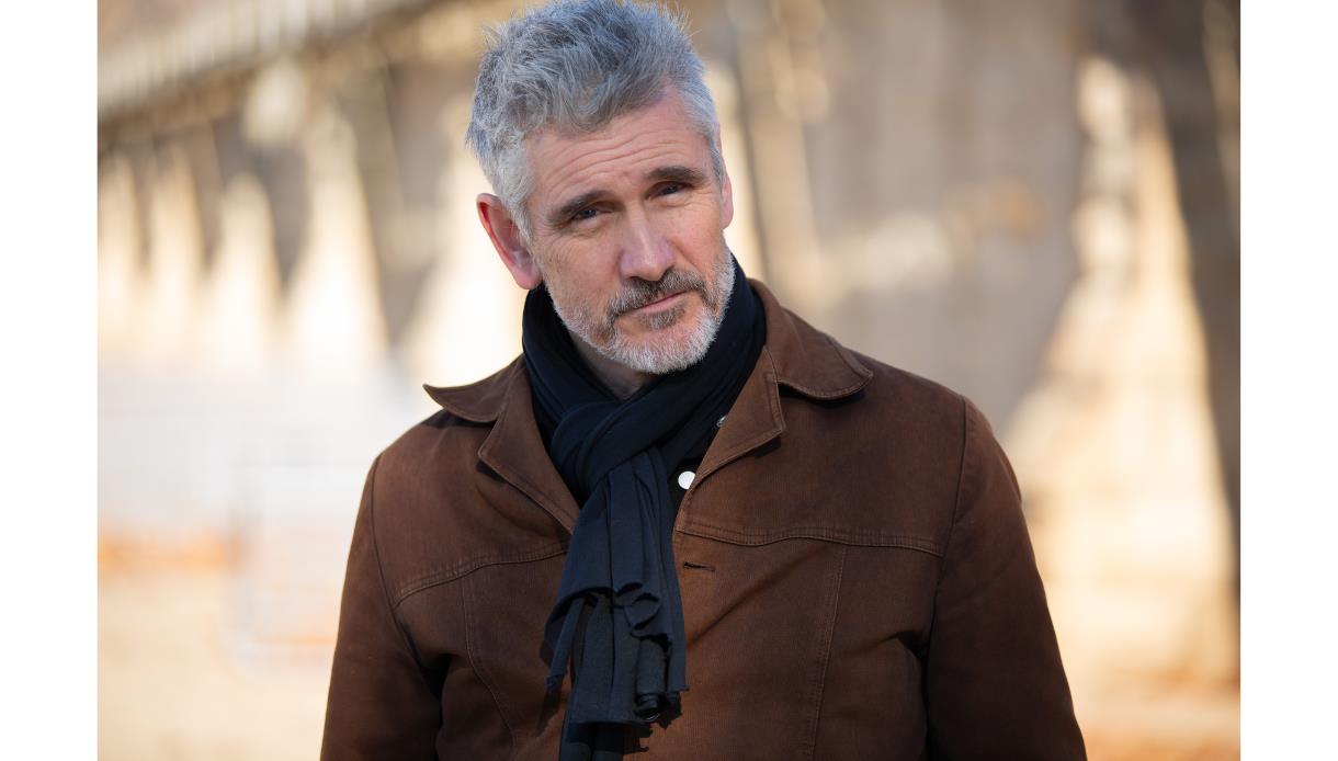 Portrait photo of a Curtis Stigers in a brown jacket and black scarf, standing outdoors with soft-focus background.