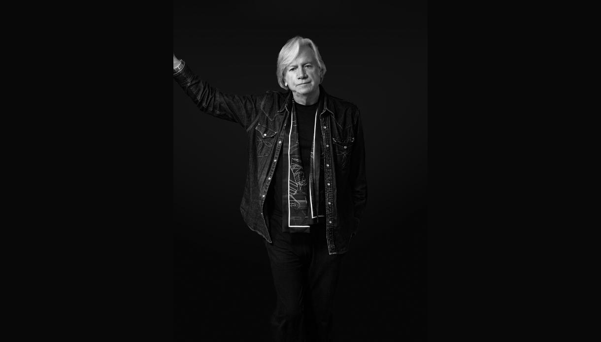 Black-and-white portrait for “The Voice of the Moody Blues: Justin Hayward,” musician with raised arm against dark background.