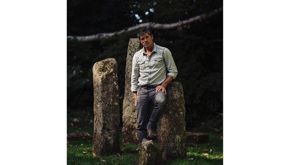 Seth Lakeman in a rolled-sleeve shirt leaning on ancient standing stones in a wooded outdoor setting.