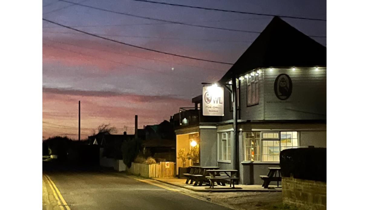 The Owl pub in Camber at dusk, with warm lights glowing and a pink sunset sky behind the quiet street.