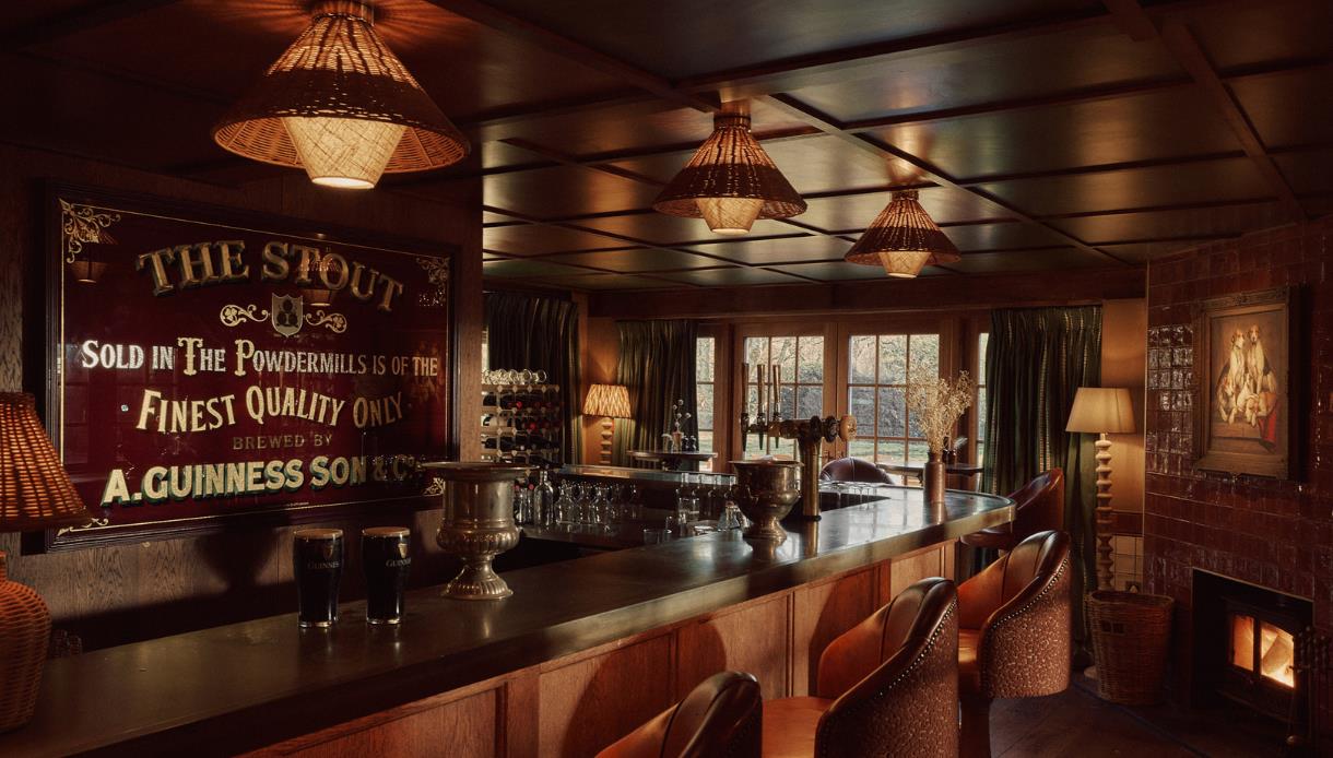 Cosy bar at Crafted at Powdermills with wooden counter, vintage sign, warm lighting and a lit fireplace.