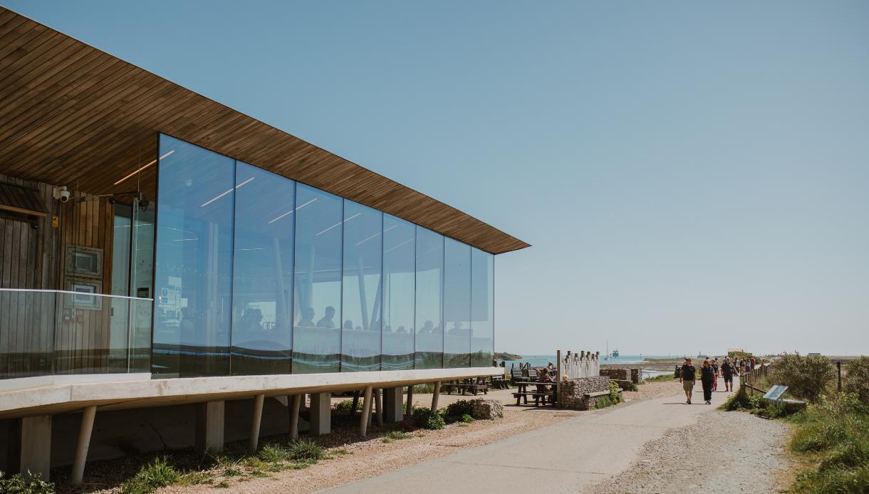 Modern glass visitor centre beside a coastal path at Rye Harbour Nature Reserve, with walkers, benches, and sea views.