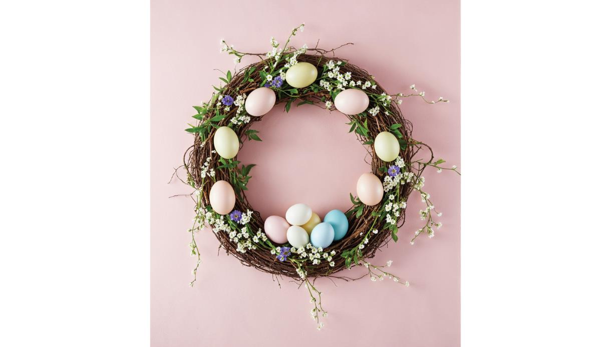 A spring wreath decorated with pastel eggs, greenery, and small flowers on a soft pink background.