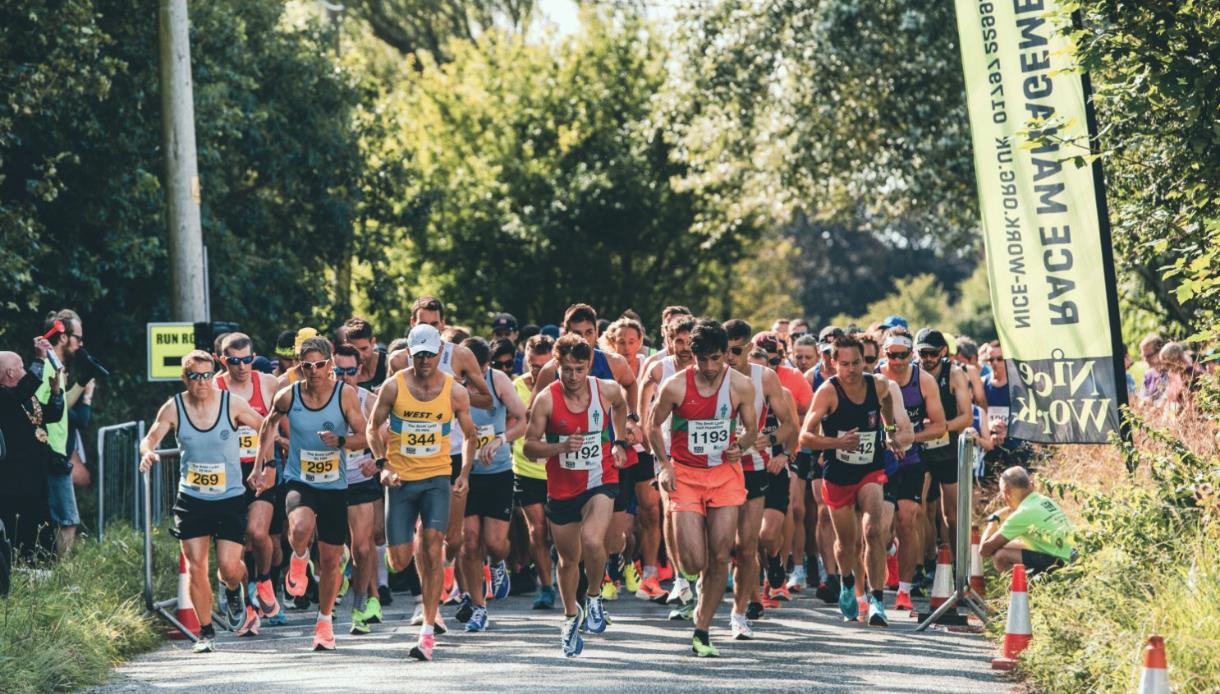 Photo of a Nice Work Race event. The photograph shows  lots of runners on the route of a race. They are all packed together and running on a road