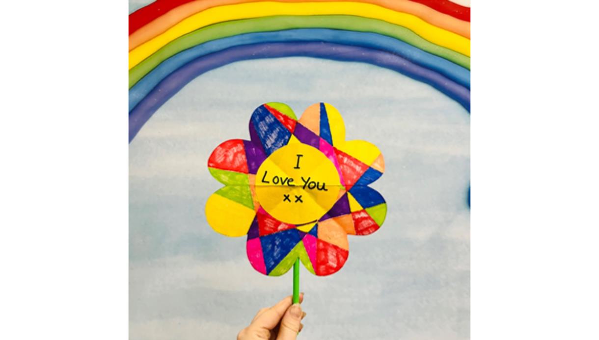 Hand holding a colourful heart-shaped pinwheel reading “I Love You xx” against a rainbow and soft blue sky background.