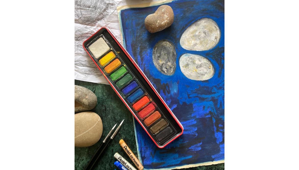 Image show a paint pallet with multiple different colours, some pastel crayons and a sketch drawn on paper