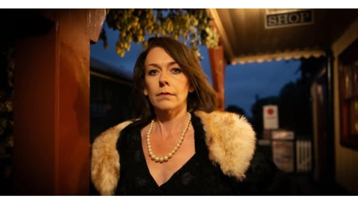 Person in vintage-style clothing with a fur stole and pearl necklace standing on a dimly lit train platform.