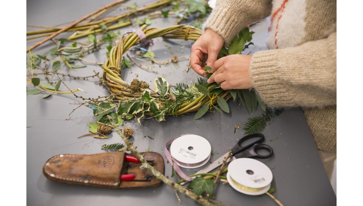 image shows someone making a christmas wreath with natural materials including leaves and holly. There are tools including scissors and ribbon.