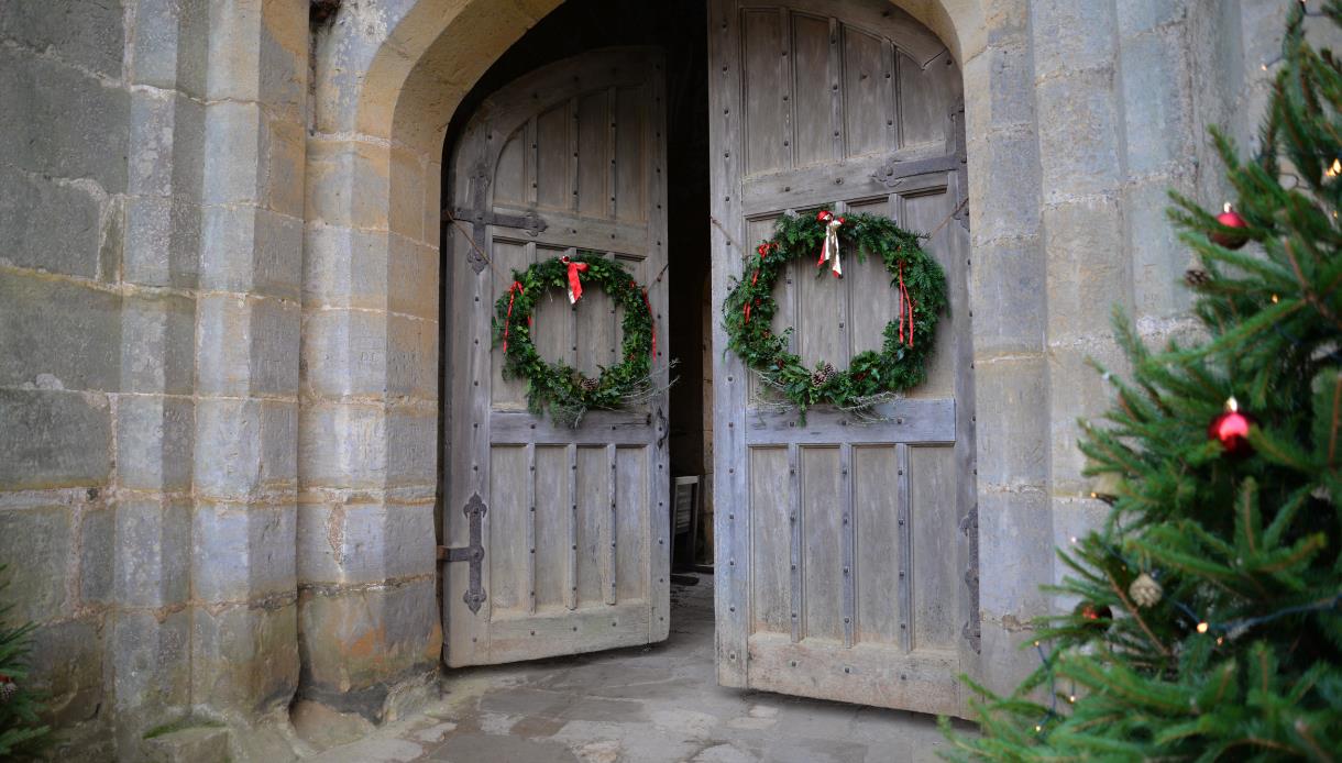 image shows two doors at bodiam castle with christmas wreaths on. In the corner of the image, you can see part of a christmas tree with red baubles on