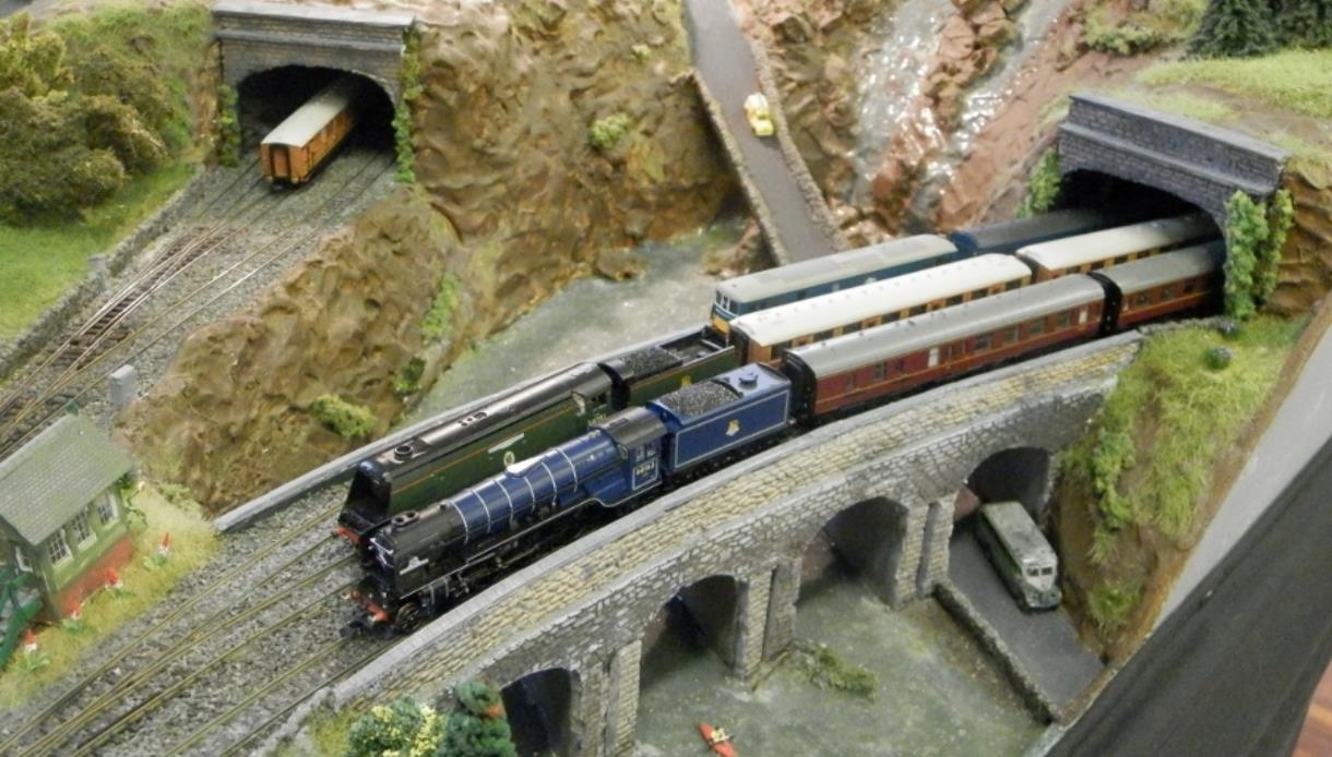 Model railway scene featuring a detailed steam train crossing a stone viaduct between two tunnels in a rocky landscape.
