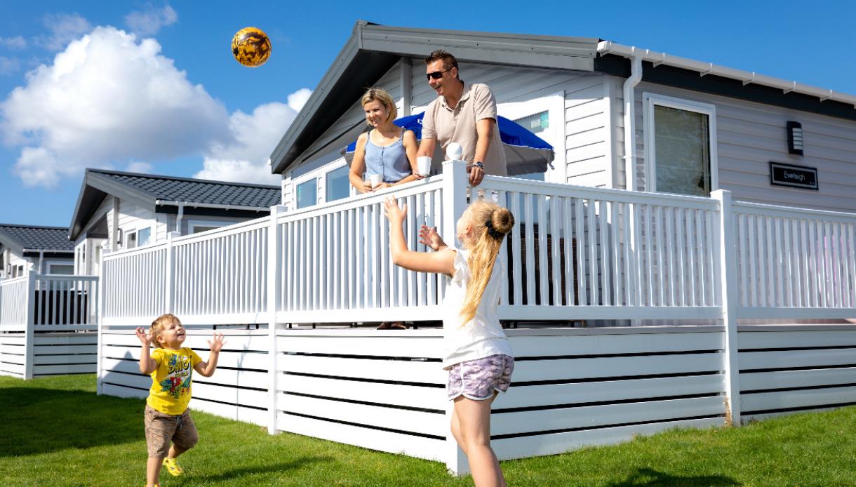 Image shows a caravan at Pevensey with 2 adults on the decking and 2 children playing a ball game on the grass