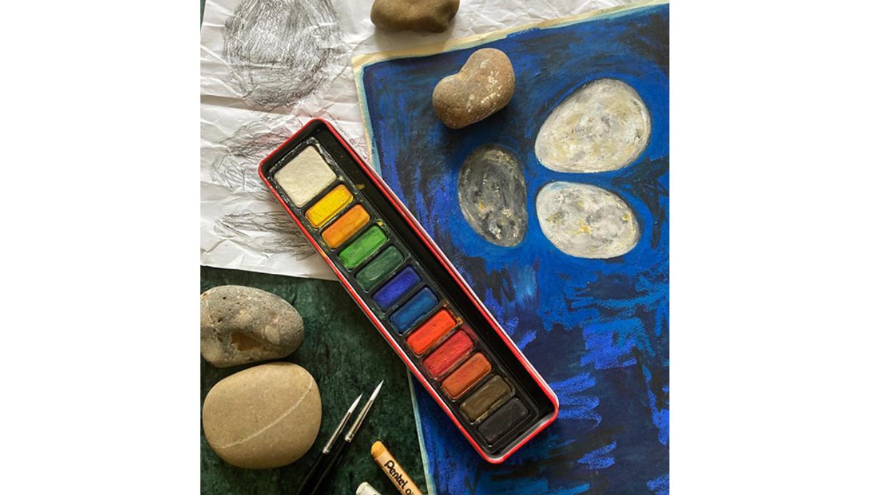 Art workspace scene featuring watercolours and brushes beside a cloudy water cup and several rounded grey stones on a tabletop; warm lighting and shal