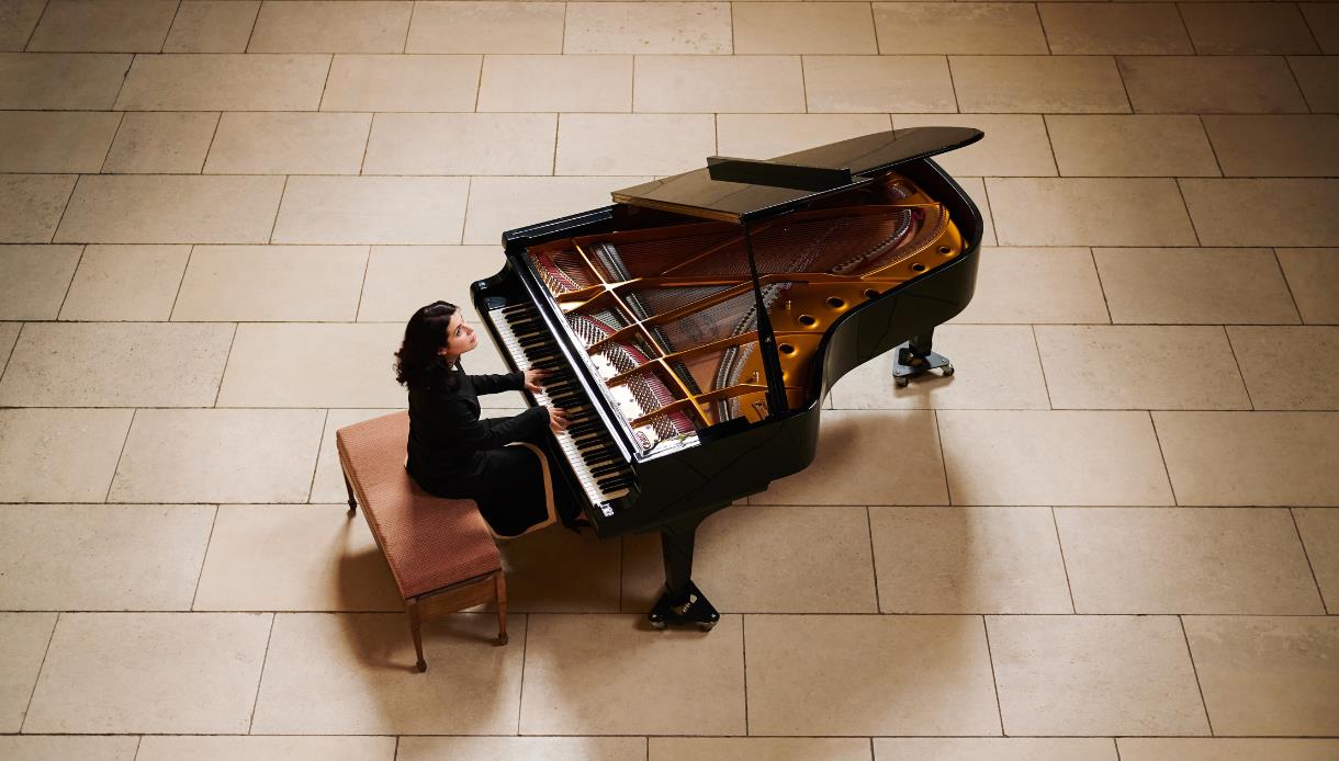 Person seated at a grand piano in a large tiled hall, playing with the piano lid open.