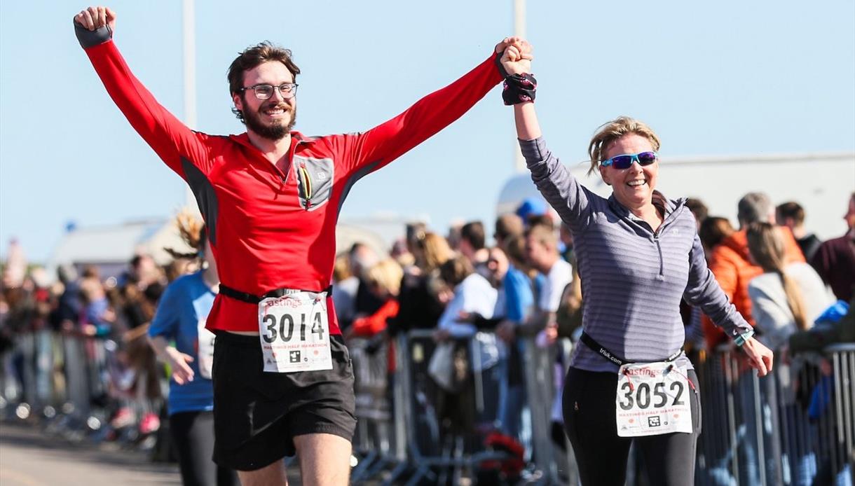 Two runners holding hands cross the finish at the Hastings Half Marathon, wearing race bibs as spectators line the course.