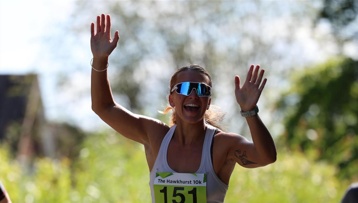 Runner wearing a numbered bib raises both hands while finishing a 10k race outdoors on a sunny day.