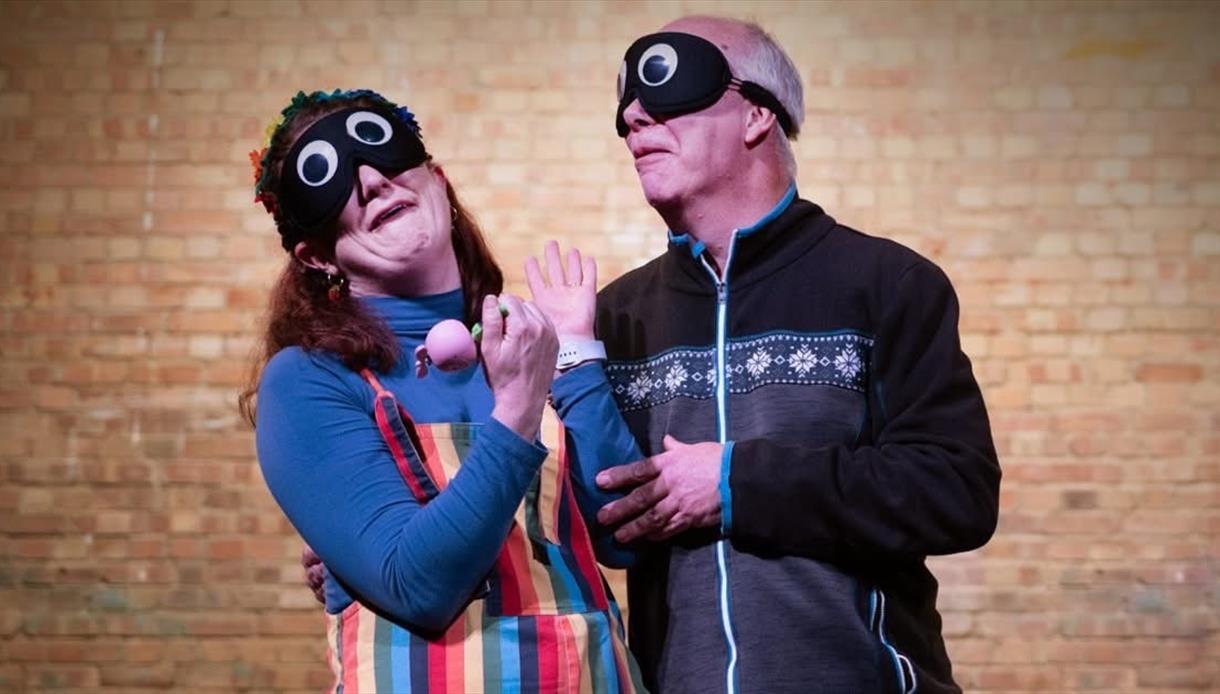 Two performers wearing playful eye masks act together on stage against a brick wall.
