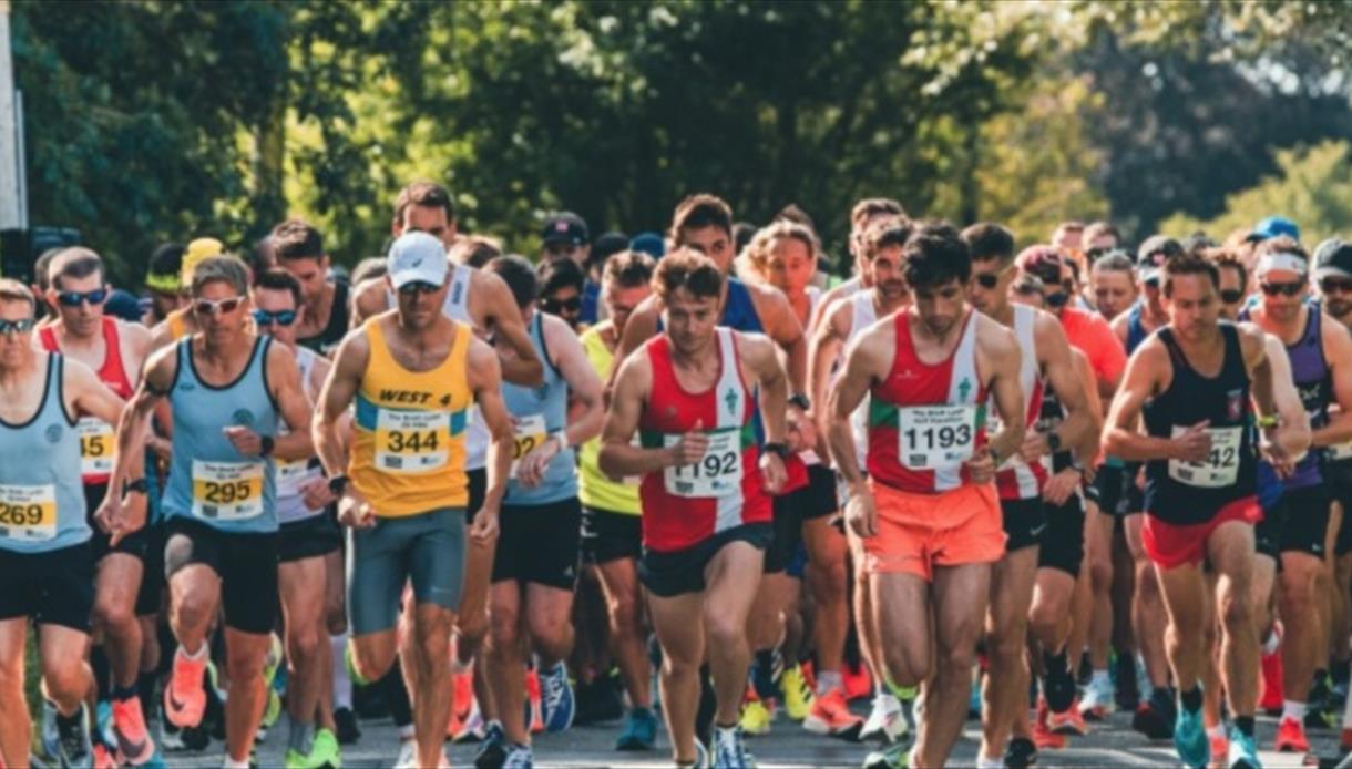 Large group of runners in athletic gear starting a race on a sunny outdoor road lined with trees.