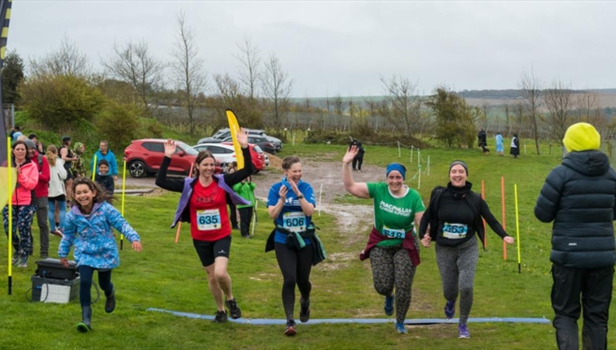 Five runners crossing a finish line on a grassy outdoor course with spectators and cars in the background.