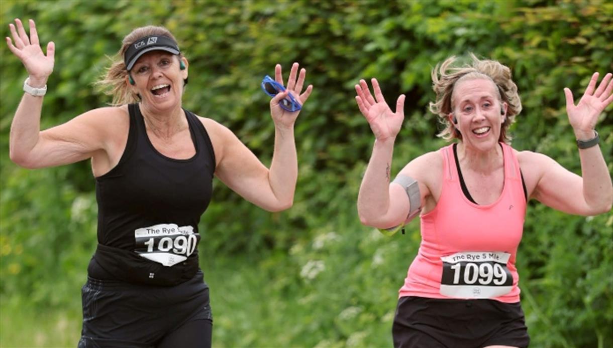 Two runners with race bibs raising their arms while running outdoors on a green, leafy path.