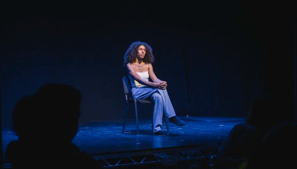 Speaker seated on a chair under blue stage lighting at “Trans Art and Activism: How do we shape the world?” event.