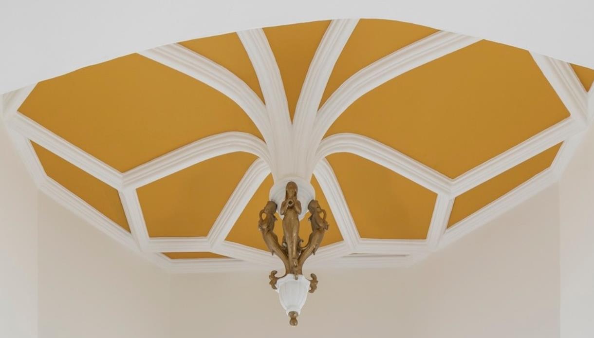 Ornate ceiling with white geometric beams and a gold background, featuring a decorative hanging light.