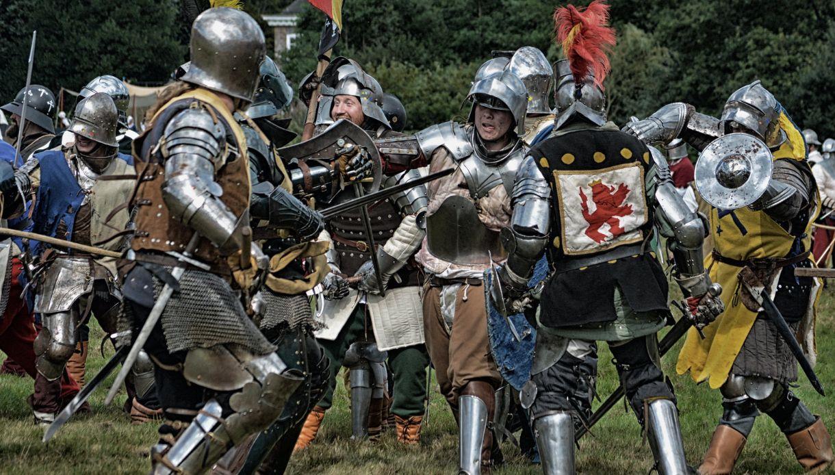 Armoured fighters in a medieval battle re-enactment clashing with swords and shields on a grassy field.