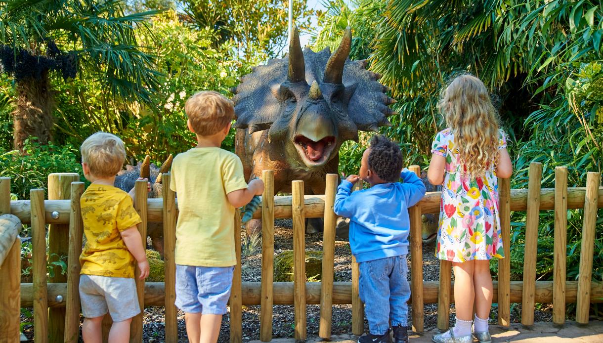 Children standing by a wooden fence looking at a large dinosaur model at Drusillas Park.
