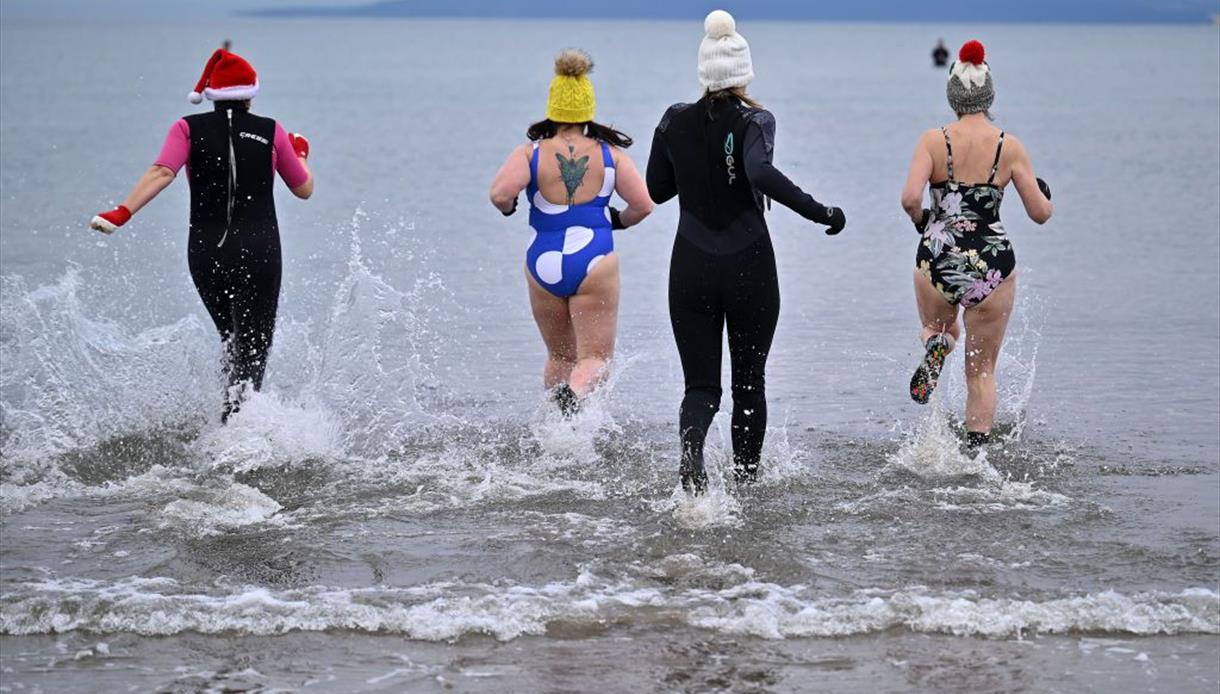 Four people are running into the sea from a sandy shoreline, creating splashes as they enter the water. They are dressed in festive and winter-themed