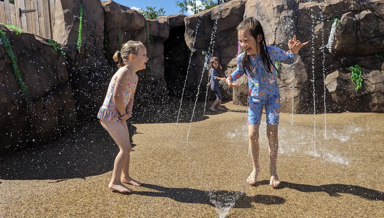 Children playing and running through water jets at Knockhatch in an outdoor splash area surrounded by rocky walls.