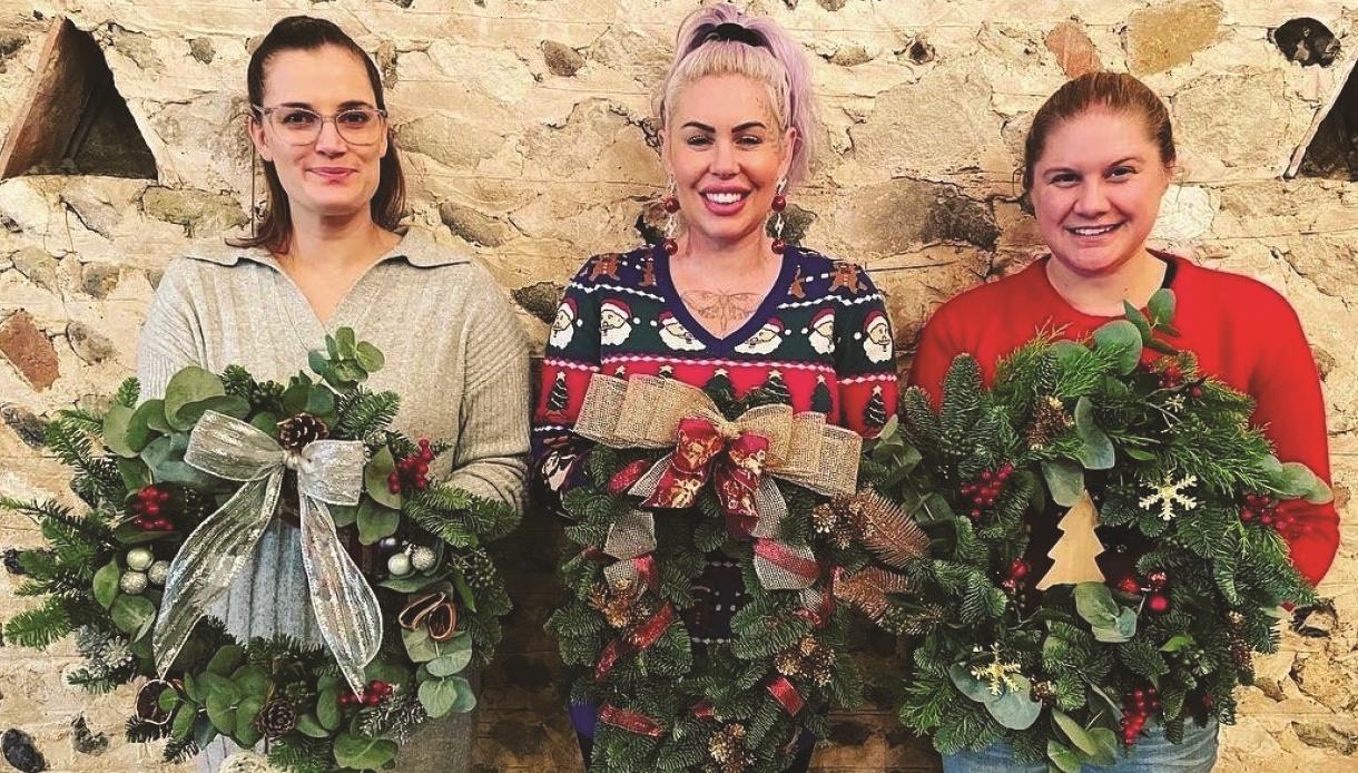 Three women hold up Christmas wreaths.