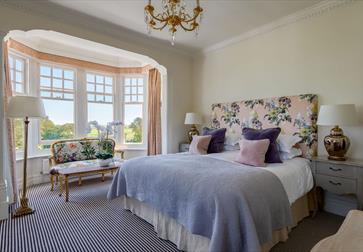 an interior photograph of a hotel bedroom at rye lodge. shows double bed with floral headrest. mauve throw, and bay windows.