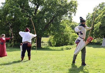 Two men and women extending bows and shooting archery targets on lawn. They are in medieval dress.