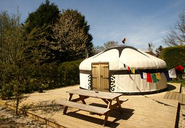 A yurt on decking with a bench in front. There is blue sky and bunting.