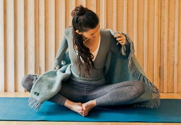 Image shows a person sitting on a yoga mat with their legs bent and feet together. They are wearing leggings, a jumper and putting a blanket around th