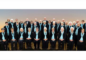 Panoramic group shot of an adult choir: approximately 35–40 people in black attire with light blue scarves, arranged in four neat rows. Front row seat