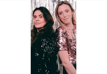 Two people wearing sparkly black and rose-gold sequin tops standing in front of a silver tinsel backdrop.