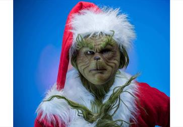 Image shows a picture of 'The Grinch' with green skin wearing a santa outfit which is red with white detail. The background of the image is blue.
