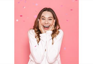 A young woman stands against a bright pink background, smiling widely with her eyes slightly closed and hands pressed to her cheeks in excitement. She