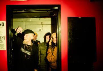 A dimly lit indoor scene with four people standing inside a doorway framed by a black doorframe. The surrounding wall is painted bright red, and there