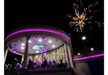 Image shows a band stand with a band playing on it surrounded by purple lights. There is a firework going off in the sky in the background.