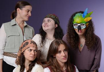 Studio photo of five costumed performers. The rear row features a smiling person in a light utility vest, a person with a patterned headscarf smiling