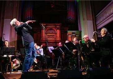 Musician performing energetically on stage with a live band in a grand hall featuring ornate wooden panels and organ pipes.