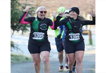Action shot of two women competing in an outdoor race, focused and energetic, with crowd barriers and city street in the background.