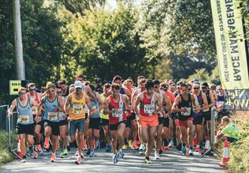 Photo of a Nice Work Race event. The photograph shows  lots of runners on the route of a race. They are all packed together and running on a road