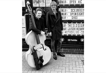 Two people standing outside a venue with a large double bass, next to a sign listing upcoming music events.