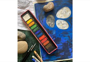 Image show a paint pallet with multiple different colours, some pastel crayons and a sketch drawn on paper